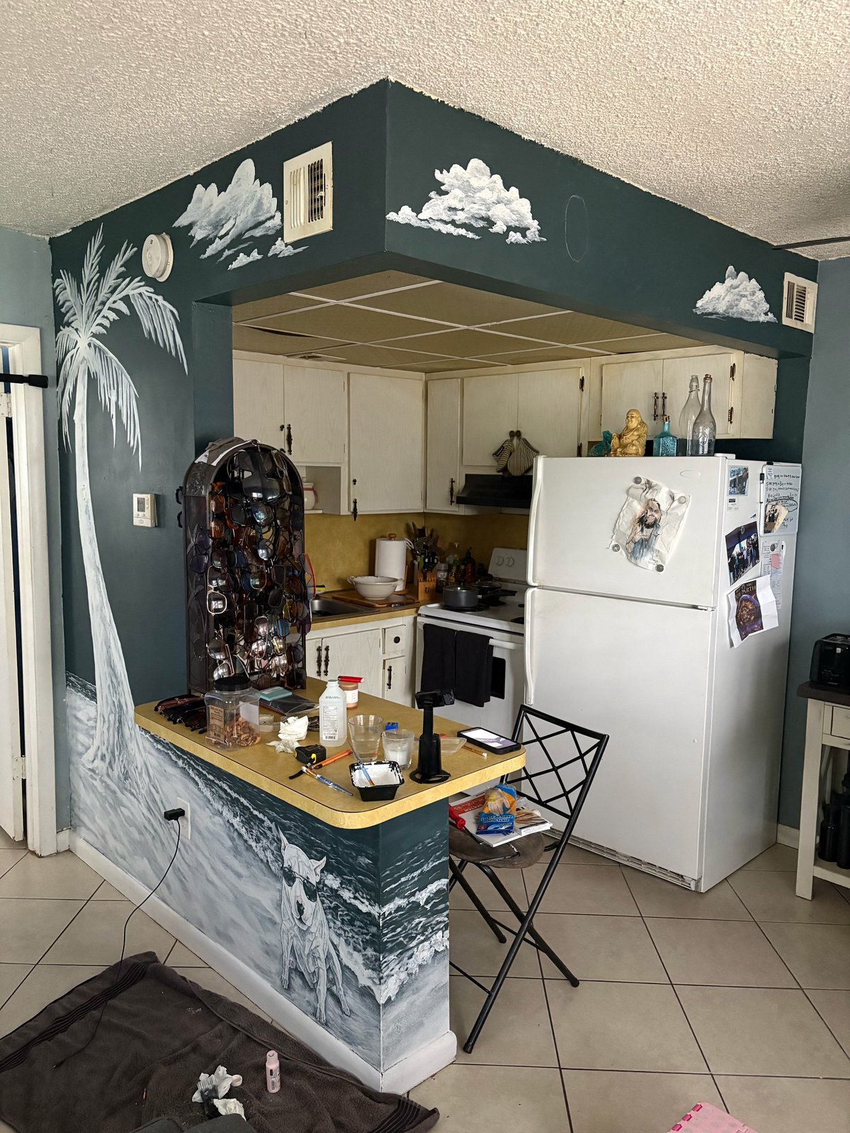 A small kitchen with dark green upper cabinets decorated with painted clouds and palm tree designs, featuring white appliances and a wooden counter with bar seating