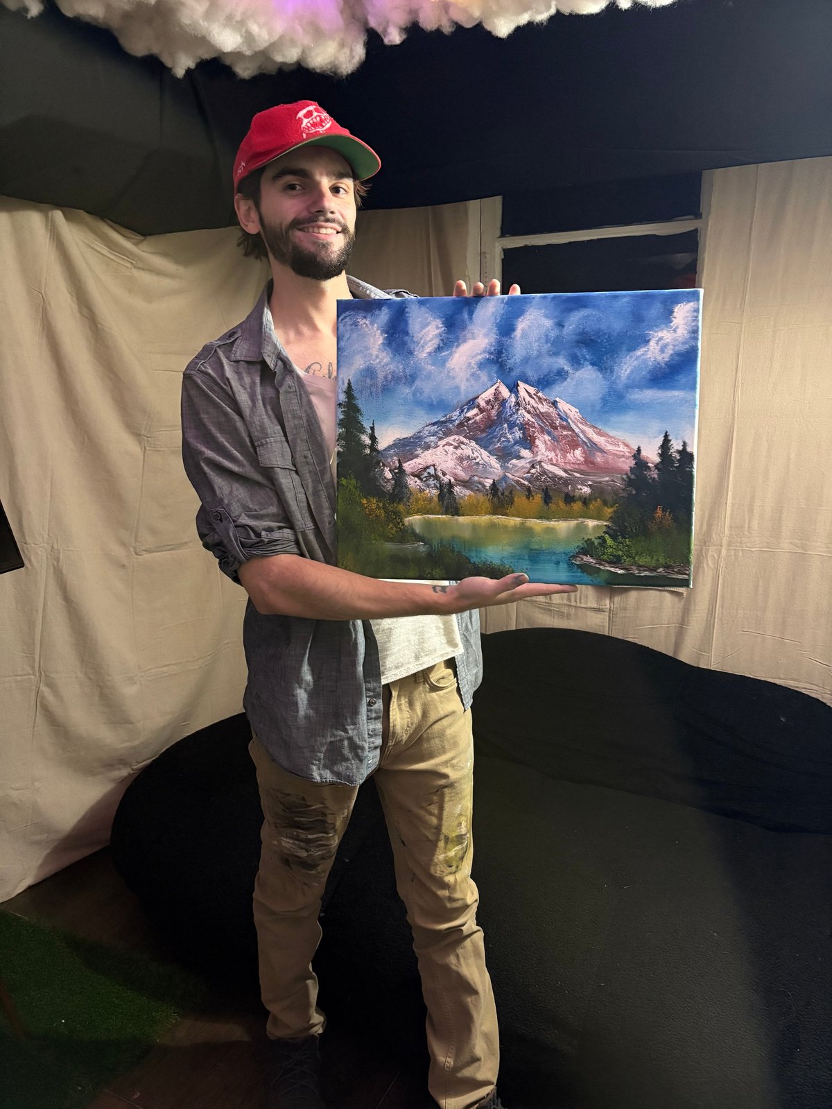 Man wearing red cap holding landscape painting of mountain lake surrounded by trees indoors