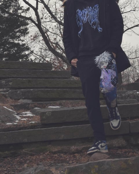Person wearing black metal band hoodie and anime character graphic on pants, standing on wooden logs in autumn forest setting