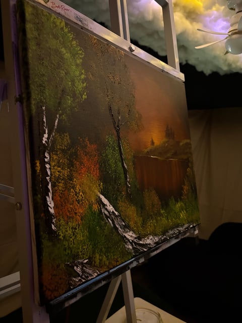 Landscape painting displayed on an easel showing birch trees in a forest with warm autumn colors and atmospheric sky