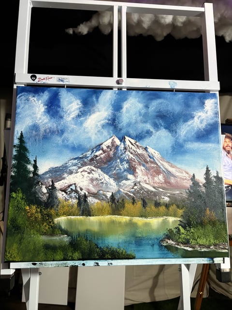 Oil painting of snow-capped mountain with forested shoreline and blue lake, displayed in white frame