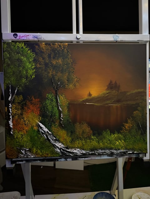 Oil painting of a serene lake at sunset framed by birch trees and autumn foliage on an easel