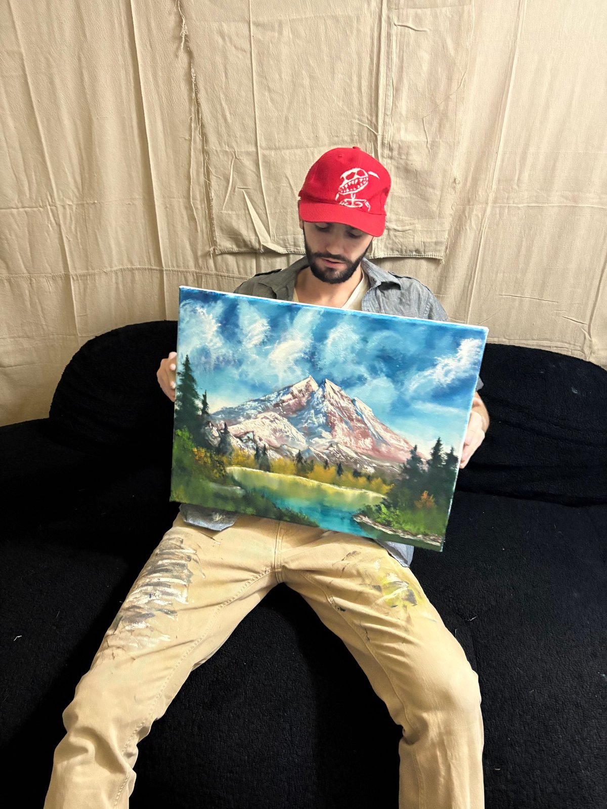 Man wearing red cap sitting on black couch holding landscape painting of snow-capped mountain by lake