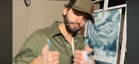 Man wearing olive green shirt and khaki hat giving double thumbs up gesture indoors
