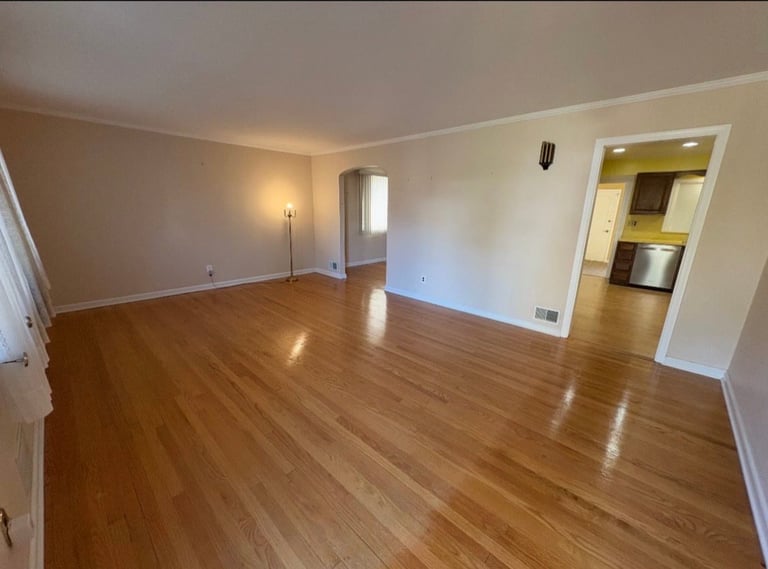 Empty apartment living room with wood flooring, white walls, floor lamp, and doorway to kitchen
