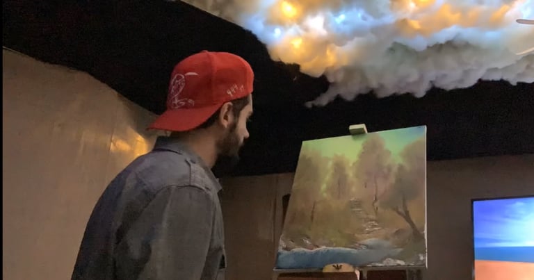 Person wearing red cap viewing landscape artwork in gallery with dramatic sky display and additional screens visible