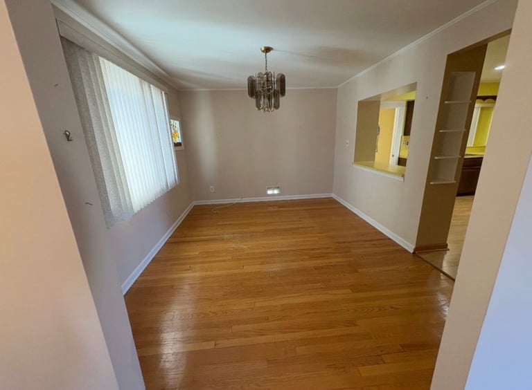 Empty living room with wooden floors, white walls, pendant light, windows with vertical blinds, and doorways leading to other rooms