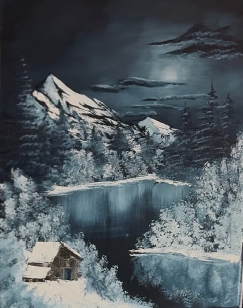Snow-covered cabin beside a frozen lake with snow-laden evergreen trees and snow-capped mountains under moonlit night sky